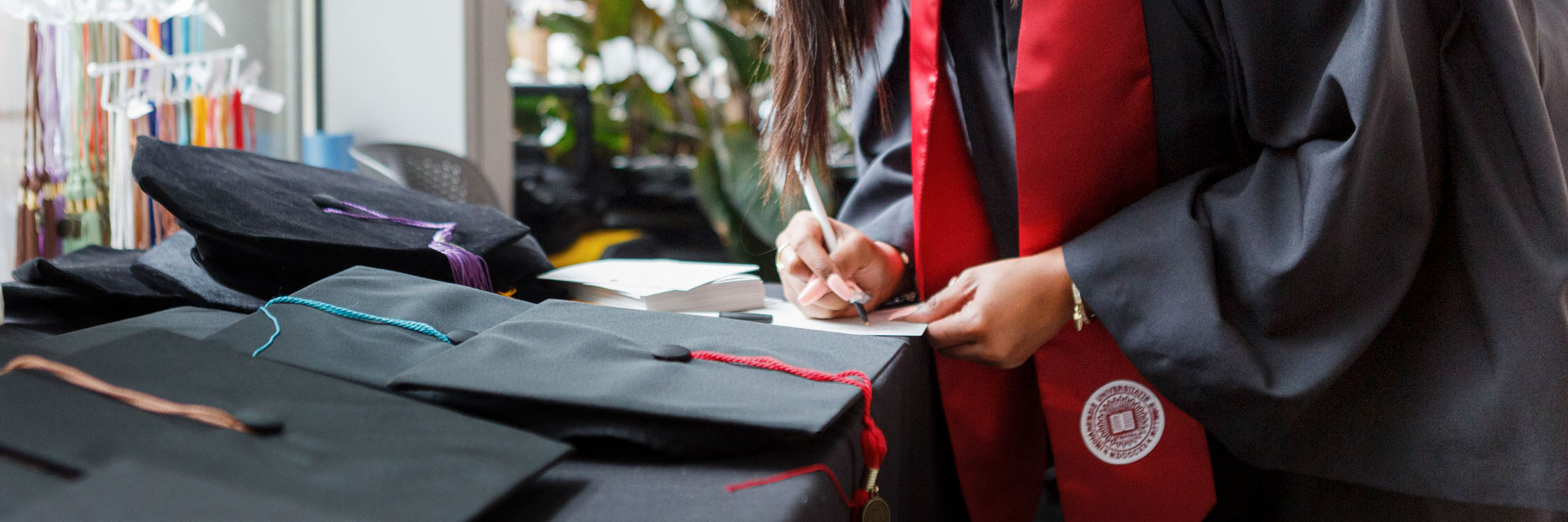 A female graduate in robes and IU crimson stole fills out paperwork behind a table full of graduation caps
