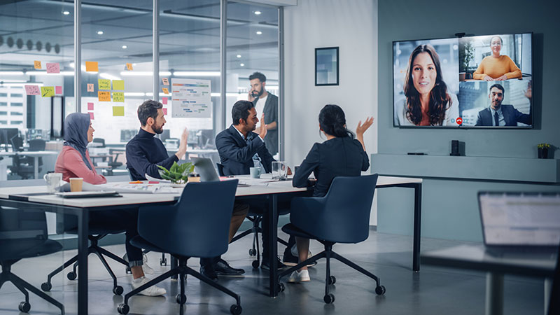 Diverse group of employees in a conference room waving at a monitor during a Zoom call