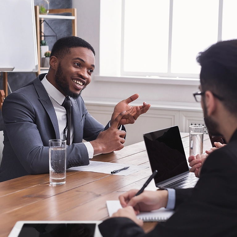 Black man in a suit gesturing toward two men on the opposite side of the table in a meeting.