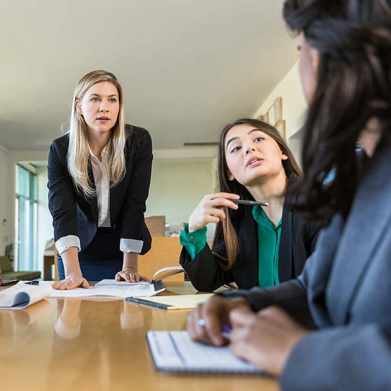 A blonde woman standing at a table where two dark haired women are seated at a meeting.