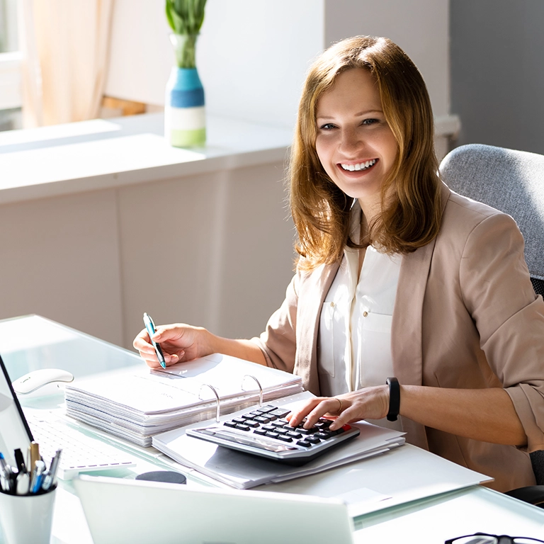 Woman with red hair wearing a white shirt and tan jacket sitting at a desk with an open binder and a calculator.