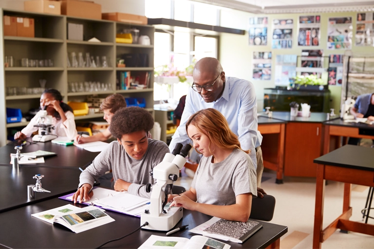 Science teacher leaning over two students' desks as they look through a microscope and take notes