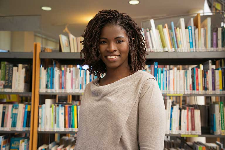Graduate student with braids smiling and standing in front of a bookcase in a library